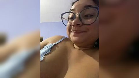 Media: Video of a plus-sized Latina woman with light brown skin, wearing glasses, a lacy bra, and a necklace, smiling, indoors.