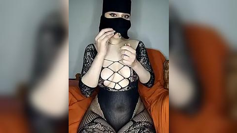 Media: A video of a woman in a black lace bodysuit with a black mask, seated on an orange couch, with a blurred background.