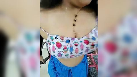 Media: A video of a light-skinned woman with medium breasts wearing a colorful, patterned crop top and blue pants. The background is blurred.