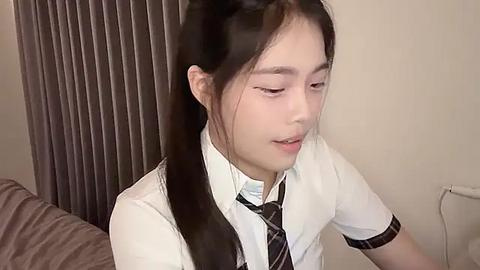 Media: Video of an Asian girl with straight, dark hair tied in a ponytail, wearing a white school uniform with a striped tie, sitting in a beige room with a gray curtain.