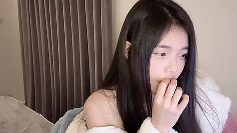 Media: A video of an East Asian woman with long black hair, wearing a white off-shoulder top, sitting on a bed, covering her mouth with her hand, against beige curtains.