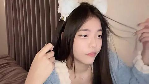 Media: Video of an East Asian woman with straight black hair, wearing white cat ear headband, light blue and white knitted cardigan, adjusting hair in a cozy bedroom with brown curtains.