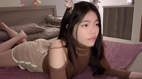 Media: Video of an Asian woman with long black hair, wearing a brown off-shoulder top and shorts, lying on a purple carpeted bedroom floor with a messy bed and gray bedding in the background.