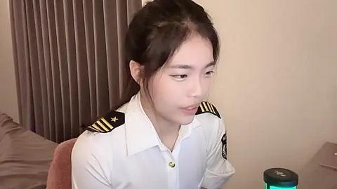 Media: Video of a young Asian woman with straight black hair, wearing a white pilot uniform with gold epaulettes, sitting on a brown chair against beige curtains.