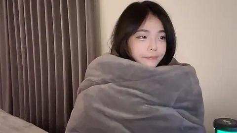 Media: Video of an Asian woman with long black hair, wrapped in a gray blanket, sitting in a dimly lit room with beige walls and vertical blinds.