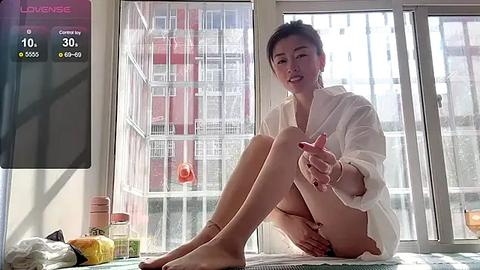 Media: Video of an Asian woman with short black hair, wearing a white robe, sitting on a bed in a bright room with large windows. Background shows a cityscape with buildings and a digital thermometer displaying 30.2\u00b0C.