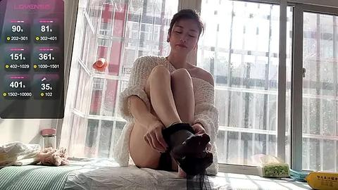 Media: Video of a young Asian woman in a white knitted sweater, sitting on a bed, putting on black high heels. Sunlight streams through a window, illuminating the room.