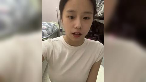 Media: Video of an East Asian girl with light skin, dark hair in pigtails, wearing a white sleeveless t-shirt, looking confused or curious, blurred background, indoor setting.