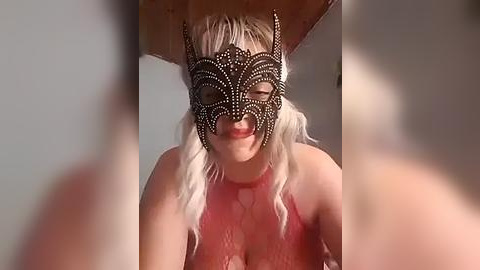 Media: Video of a young woman with platinum blonde hair, wearing a fancy black lace mask with intricate patterns, and a pink lace top, smiling. The background is blurry.