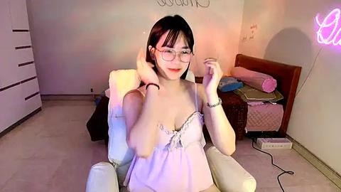 Media: Video of an Asian woman with glasses, light skin, and small breasts, wearing a lacy white camisole, adjusting her hair. She sits in a plush chair in a cozy, softly lit room with a neon sign and scattered items.