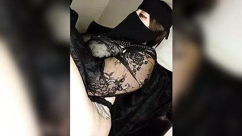 Media: Video of a person in black lace lingerie with a sheer, lace-covered bra, wearing a black mask, with blurred, shadowy figures in the background.