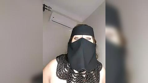 Media: Video of a woman wearing a black face mask, black headband, and a sleeveless top with white polka dots. The background features a white wall with an air conditioner mounted above.