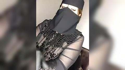 Media: A video of a woman in a black face mask, black gloves, and a sheer black dress with polka dot trim, seated indoors, with a blurred background.