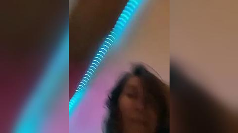 Media: A blurry video showing a person with curly hair and a blue light strip background, with a pink and beige wall. The image is slightly out of focus.