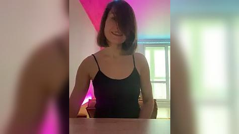 Media: Video of a smiling, light-skinned woman with shoulder-length dark hair, wearing a black tank top, in a modern, dimly-lit room with pink and blue lighting.