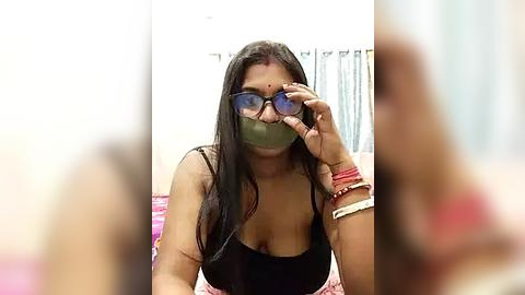 Media: Video of a young woman with long black hair, wearing a black tank top, green face mask, and glasses, adjusting them while seated on a bed with pink and purple sheets.