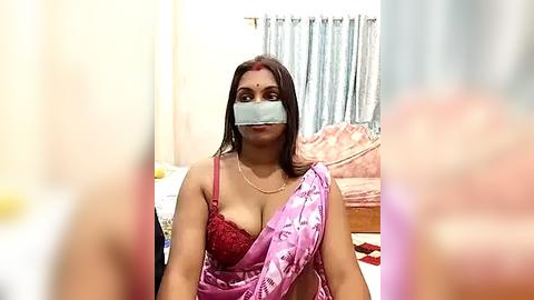 Media: Video of a woman with medium-dark skin, wearing a pink sari with red lace bra, her face covered by a gray bandana, in a cozy bedroom with floral curtains and a pink sofa.