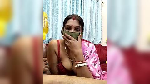 Media: Video of a South Asian woman with medium skin tone, wearing a red bra and pink sari, holding a green phone, in a cluttered room with light blue curtains.