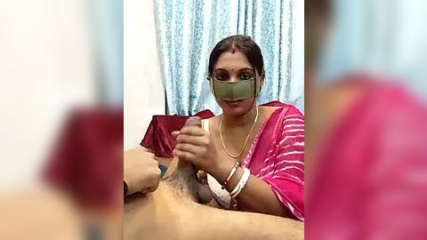 Media: Video of a South Asian woman in a pink saree, green mask, and white bracelets, holding a man's erect penis, blurred background, indoor setting.