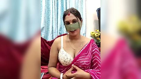 Media: Video of a woman with medium skin tone, wearing a red and white striped sari, a green face mask, and a white lace bra, sitting on a red bed.