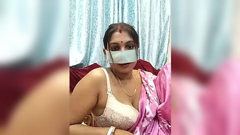 Media: A video of an Indian woman with medium brown skin and dark hair, wearing a pink saree, white bra, and a face mask, seated on a red sofa.