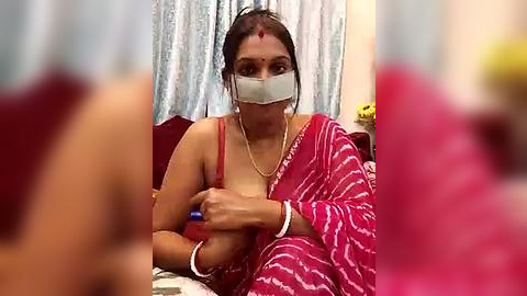 Media: Video of an Indian woman in a red saree with a white pattern, wearing a mask, sitting on a bed, holding a blue object.