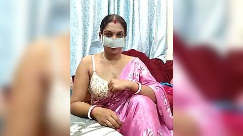 Media: Video of an Indian woman with dark skin, wearing a pink sari, white mask, and red bindi, seated on a bed with blue and white curtains, holding a phone.