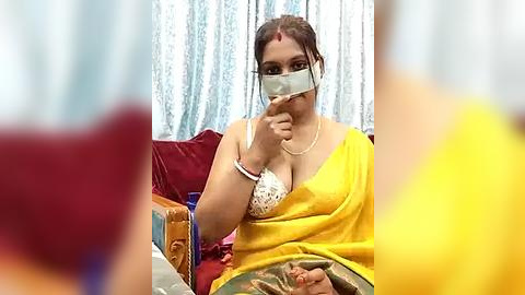 Media: Video of a woman in a yellow sari, sitting on a red couch, wearing a mask, with a blurred background.