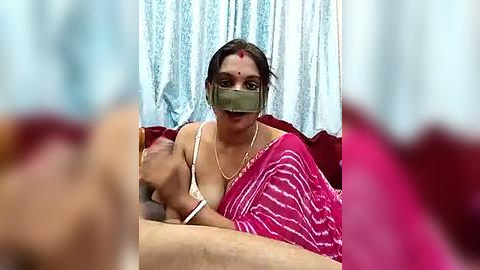 Media: Video of a woman in a pink saree with white stripes, wearing a face mask, holding a baby, in a room with white curtains.