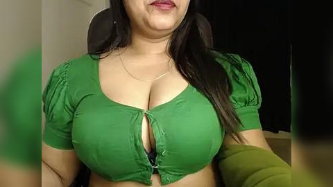 Media: Video of a woman with long black hair, fair skin, and full lips, wearing a low-cut green top that accentuates her ample breasts, against a dark background.