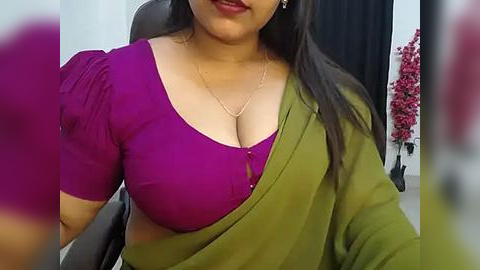 Media: Video of a South Asian woman with medium skin tone, wearing a bright purple blouse with a plunging neckline and a green sari, seated on a black office chair. Background includes a pink plant and a dark curtain.