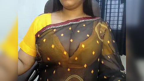 Media: Video of a woman wearing a sheer, brown saree with yellow diamond patterns and a yellow blouse, revealing her large breasts. She has a medium brown complexion and long dark hair. The background features a white wall and a partially visible black chair.