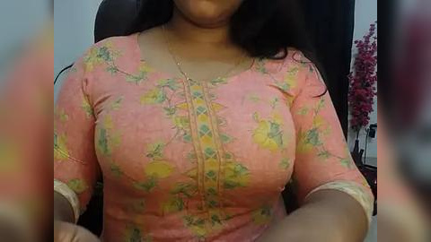 Media: Video of a woman with medium brown skin, wearing a snug, floral-patterned pink top that accentuates her medium-sized breasts. She has long, dark hair and is seated in an office chair.