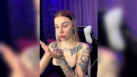 Media: Video of a tattooed, fair-skinned, blonde woman with small breasts, wearing a black tank top, sitting in a black chair with a white towel. She gestures with her hands. Background features a brick wall and purple lighting.