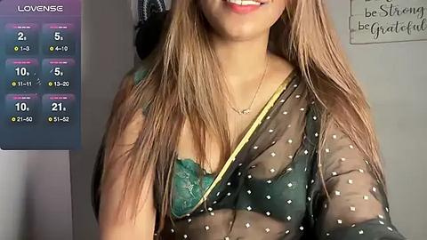 Media: Video of a smiling woman with light brown skin and long, straight brown hair, wearing a sheer black top with white polka dots, revealing a green bra underneath. Background shows a calendar and motivational quote.