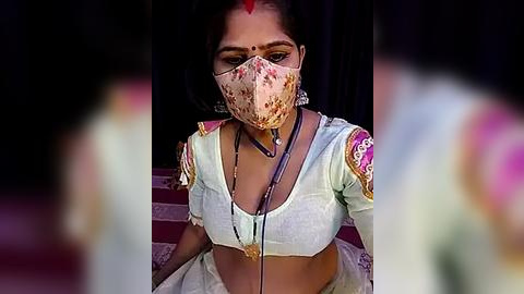 Media: Video of a South Asian woman with medium skin tone, wearing a floral face mask, light blue blouse with pink embroidery, and a pink sari, seated against a dark background.