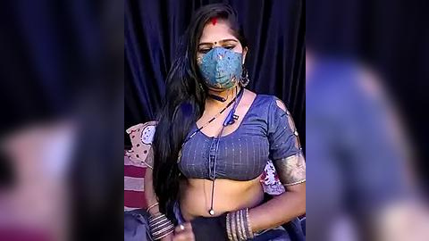 Media: Video of a woman with long black hair, wearing a blue face mask, traditional blue blouse, and gold bangles, seated against dark curtains.