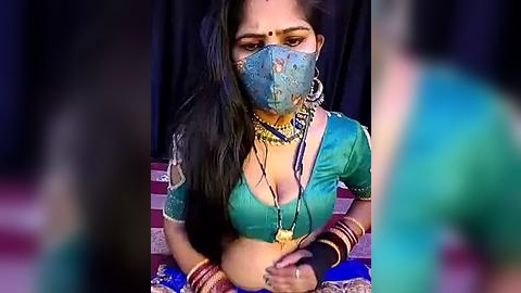 Media: Video of a South Asian woman with long black hair, wearing a blue saree, gold jewelry, and a blue mask, seated on a blue and red rug against a dark backdrop.