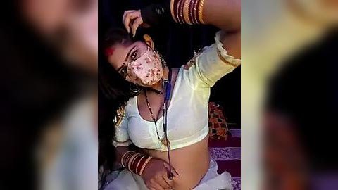 Media: Video of a dark-skinned woman with a fair complexion, wearing a white saree, gold bangles, and a face mask. She has a medium build and medium-sized breasts. Background includes blurred figures and a patterned blanket.