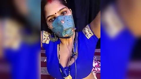 Media: Video of a South Asian woman with fair skin, dark hair, and a blue mask, wearing a traditional blue blouse with gold embroidery, sitting on a patterned red and white rug.