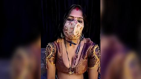 Media: A video of a woman with long black hair, wearing a floral face mask, traditional gold jewelry, and a richly patterned sari, set against a dark backdrop, exudes a serene, cultural essence.