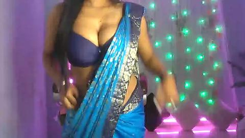 Media: Video of a woman in a blue sari with intricate gold embroidery, revealing her cleavage in a dark blue bra. She stands against a purple and green light backdrop, with a blurred, possibly dancing figure in the background.