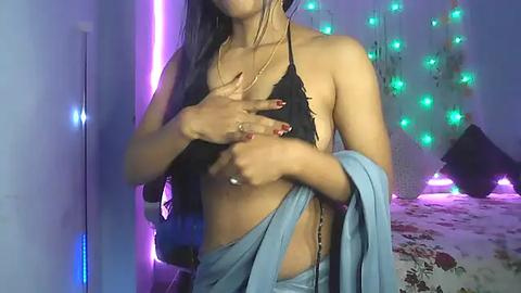 Media: Video of a slender, dark-skinned woman with long, straight hair, wearing a black halter top and light blue sarong, standing in a dimly lit room with purple and green LED lights and a bed in the background.