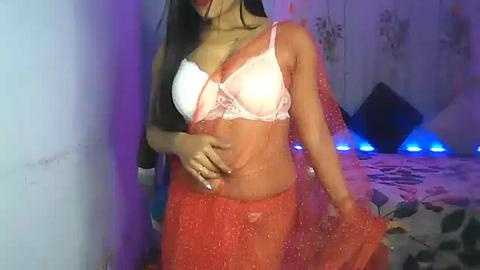 Media: Video of a South Asian woman in red saree, white lace bra, purple lighting, holding saree, on bed with pillows, indoor setting.