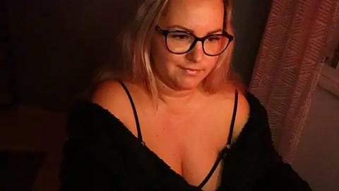 Media: Video of a fair-skinned woman with blonde hair, wearing glasses and a black lace top, revealing cleavage, sitting in a dimly lit room with a textured curtain in the background.