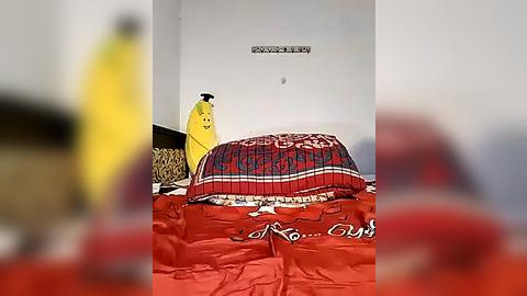 Media: A video showing a yellow banana-shaped object atop a red and white bedspread with ornate patterns. The background is blurred, with a person wearing red, partially visible.