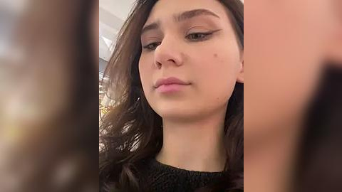 Media: Video of a young woman with fair skin and dark brown hair, wearing black clothing, focusing on her face, which has subtle makeup and a neutral expression.