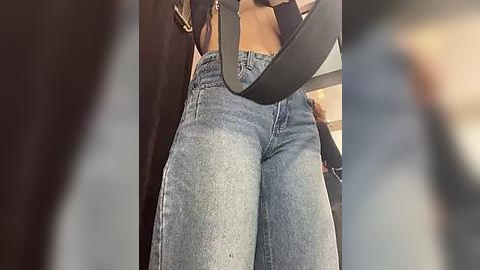 Media: Video of a woman's lower body wearing high-waisted, light blue denim jeans with a black leather belt slung diagonally across her hips. The background is blurred, suggesting an indoor setting.