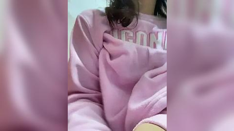 Media: Video of a person in a pink hoodie, head down, holding their face, blurred, in a soft pink and white room.