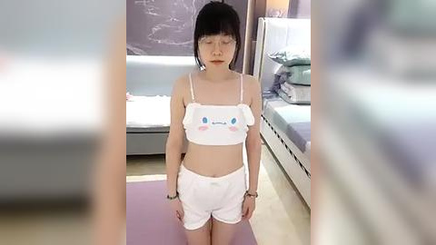 Media: Video of an East Asian woman with short black hair, wearing a white cat-themed crop top and shorts, standing on a purple mat in a minimalist bedroom with a bed and blue wall.